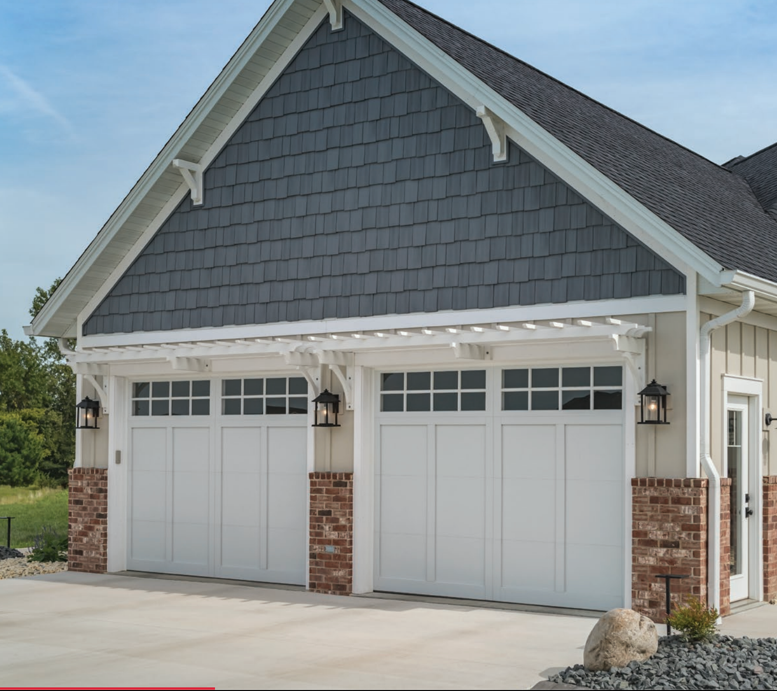 White garage doors with brick columns, under a dark blue shingled roof, and a white pergola.