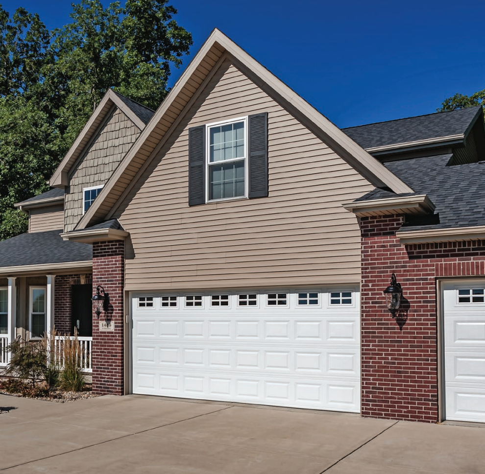 Tan and red brick home with a white garage door, blue sky, and trees.