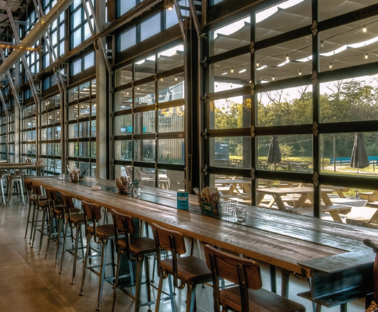 Interior of a restaurant with a long wooden table and windows overlooking an outdoor seating area.