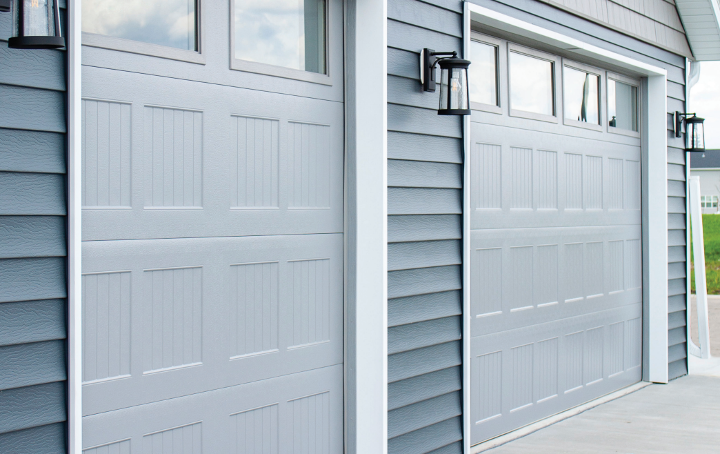 Two light gray garage doors with windows, on a house with blue siding and white trim.