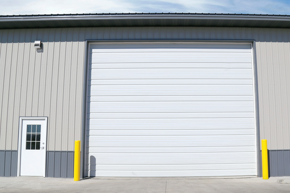 White garage door on a tan and grey building with a small white door on the left side and yellow posts on the right.