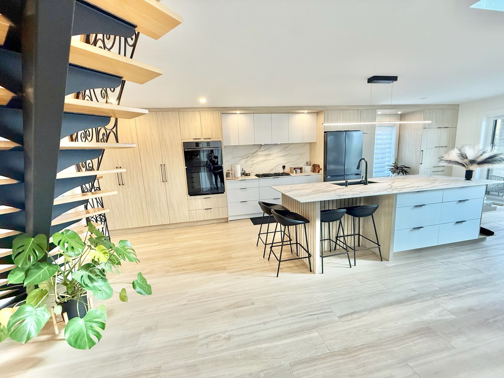 Cuisine moderne avec îlot central, coin repas et escalier en colimaçon en bois. Sol, placards et électroménager noirs en bois clair.