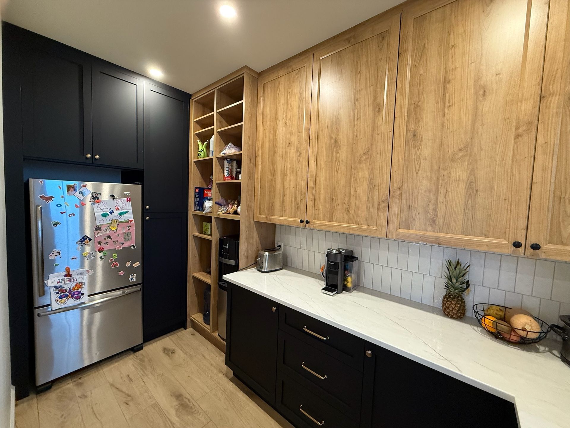 Cuisine avec placards noirs et en bois, réfrigérateur, garde-manger et plan de travail blanc.