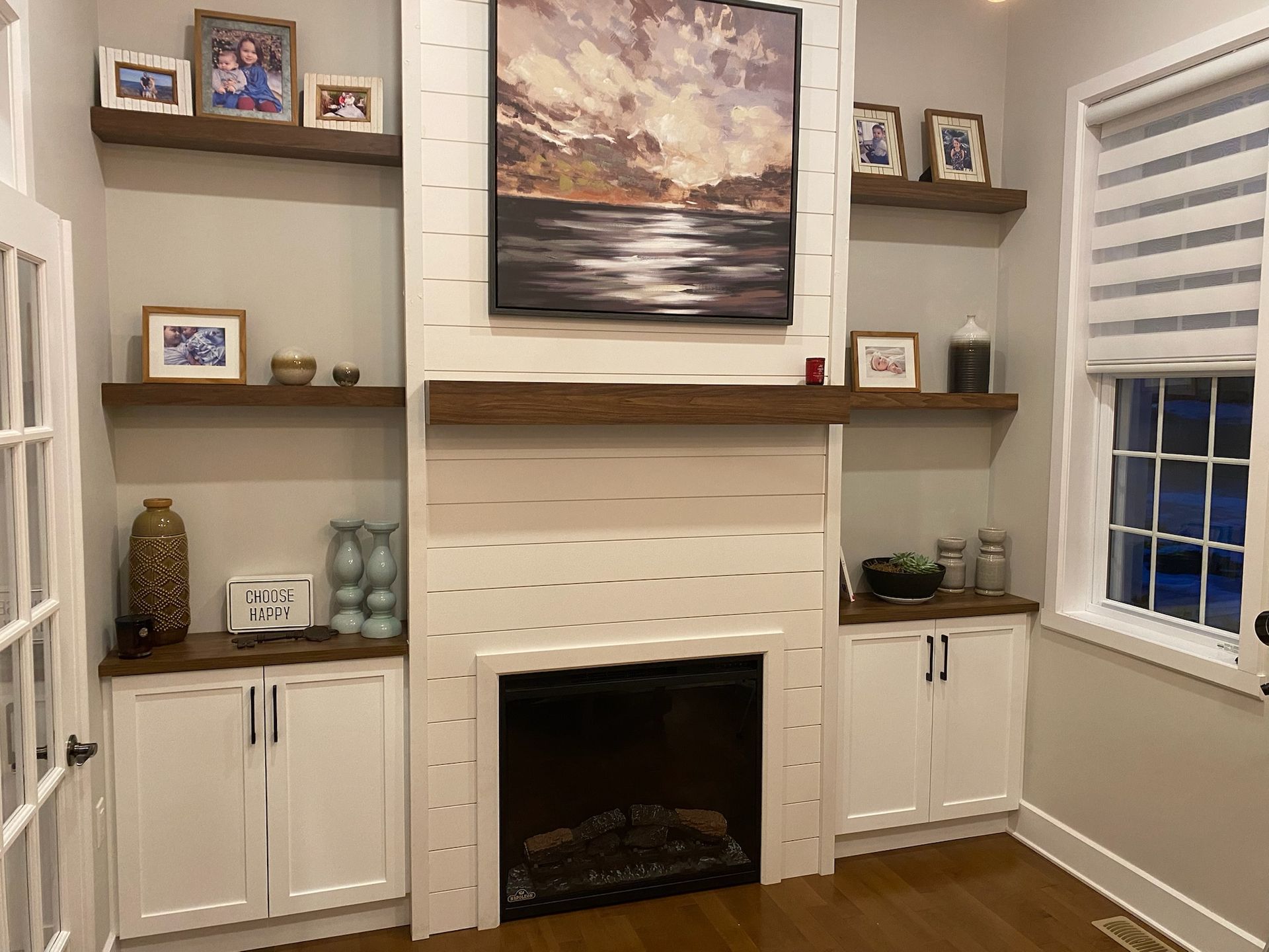 Pièce chaleureuse avec cheminée, placards blancs intégrés, étagères flottantes décorées et un grand tableau abstrait.