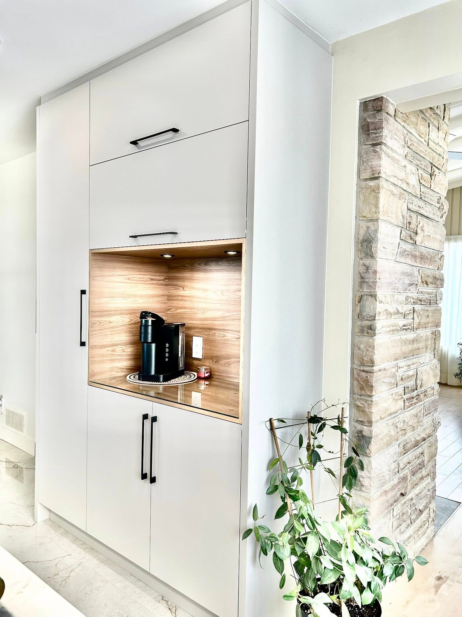 Meubles blancs intégrés avec coin café. On aperçoit des éléments en bois, une machine à café et une plante.