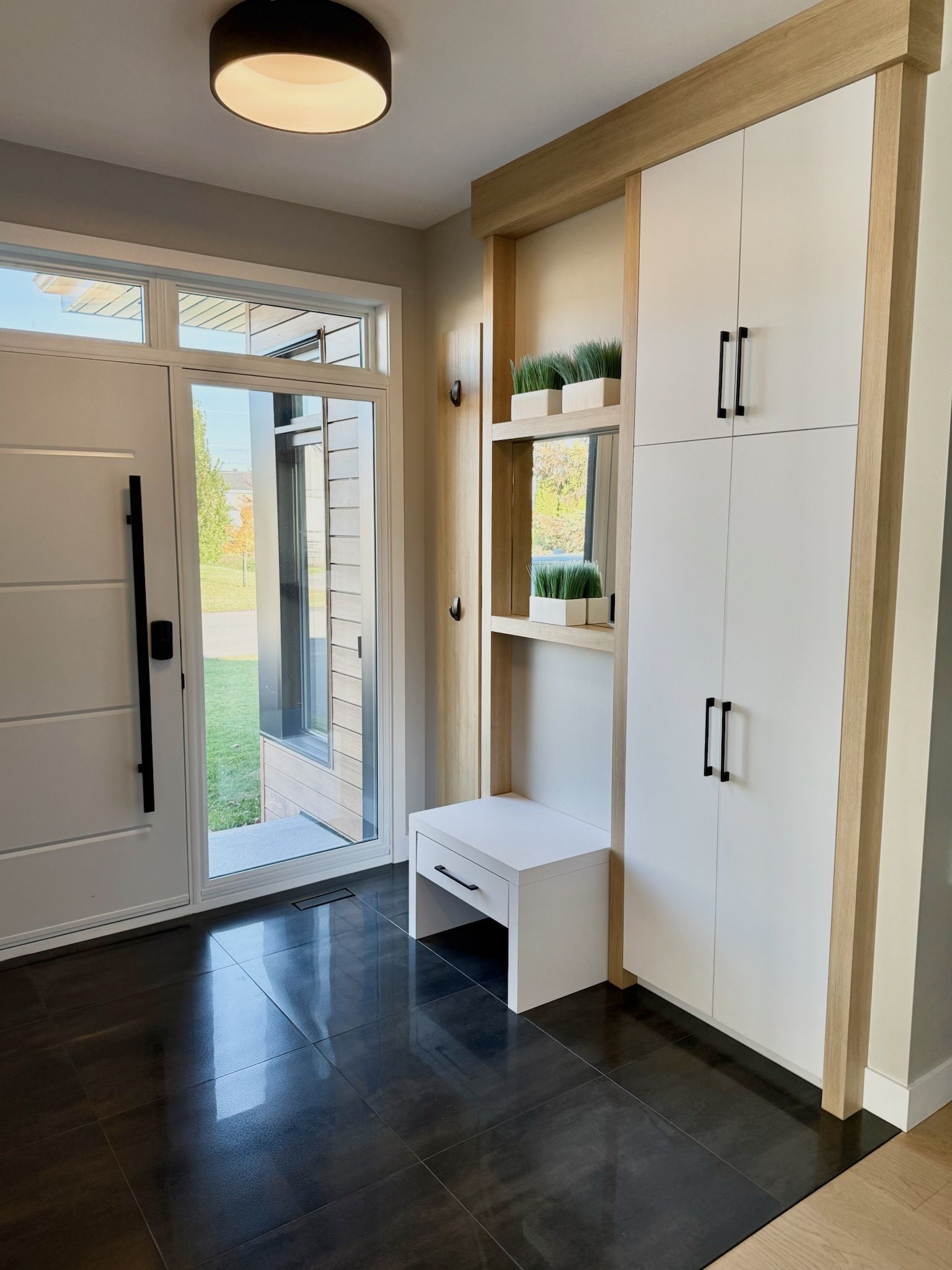 Entrée avec porte blanche, parquet, rangements intégrés, banc et plantes. Éclairage lumineux.