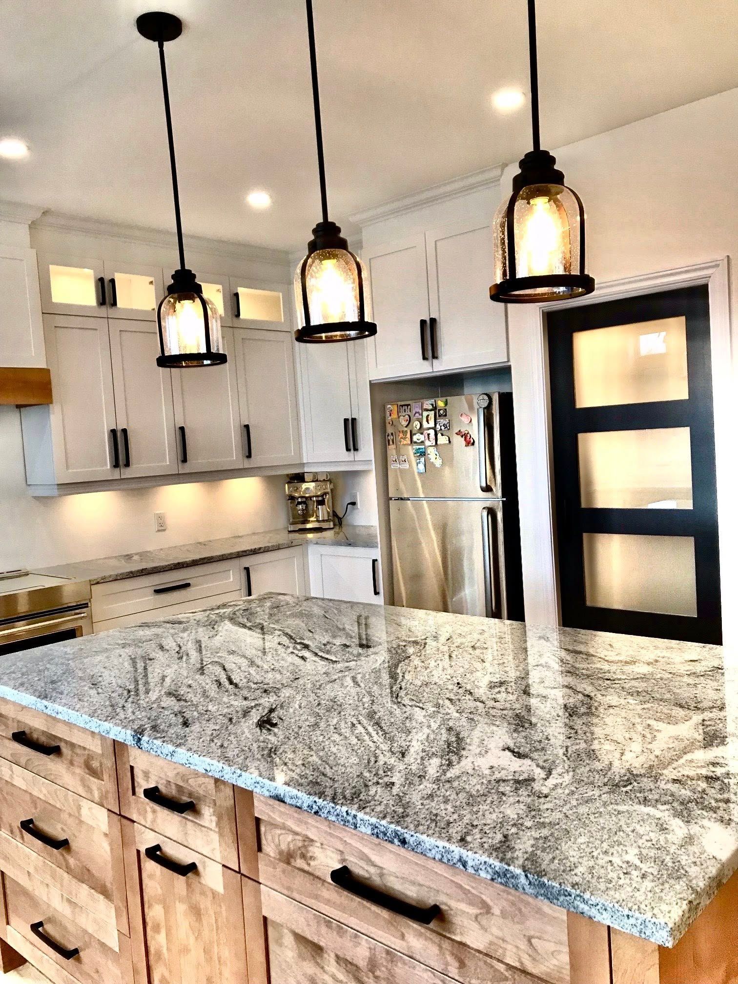 Cuisine avec îlot en granit, armoires en bois, suspensions lumineuses noires et réfrigérateur en acier inoxydable.