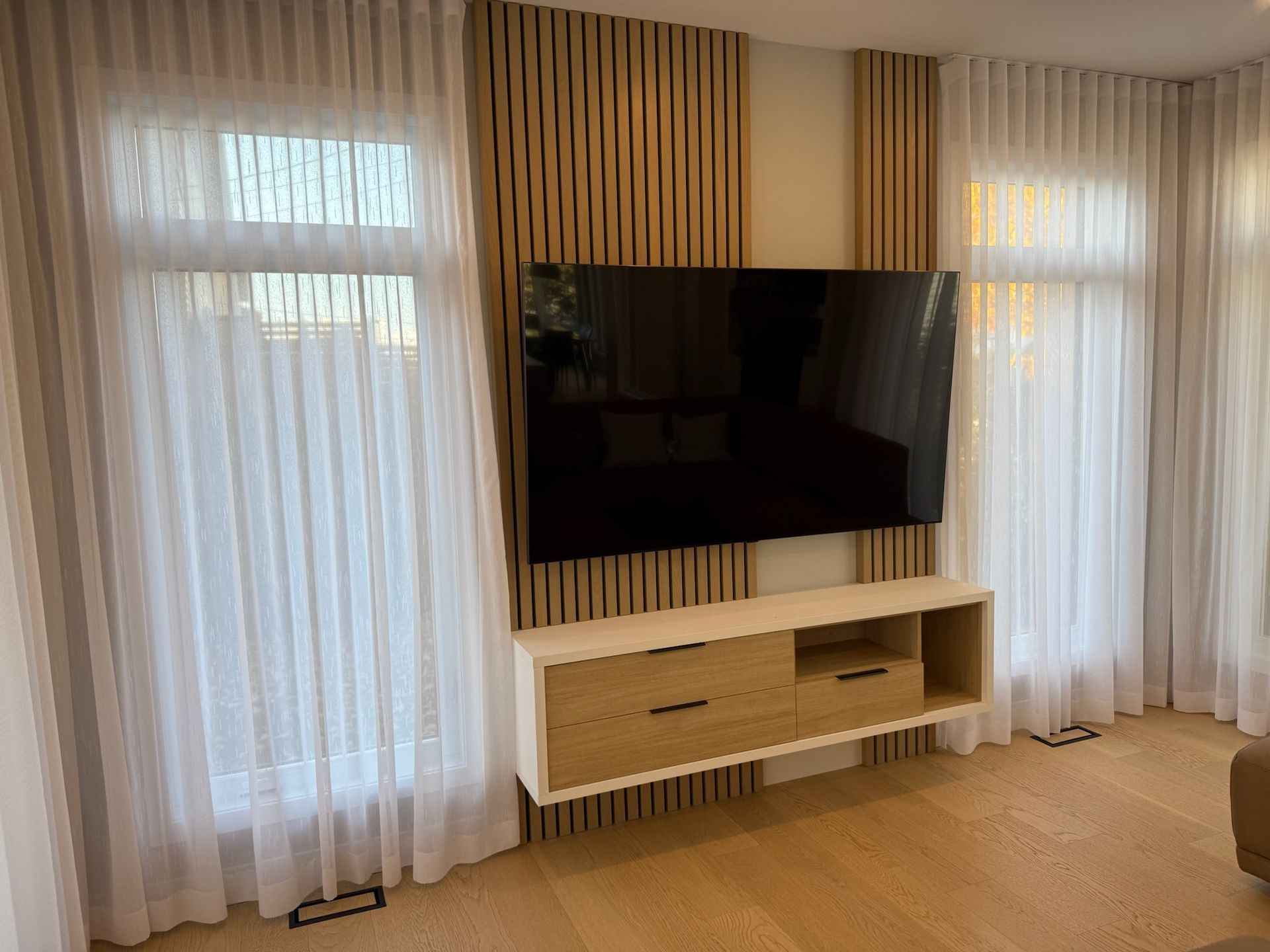 Salon avec télévision murale, lambris, meuble suspendu et voilages blancs.