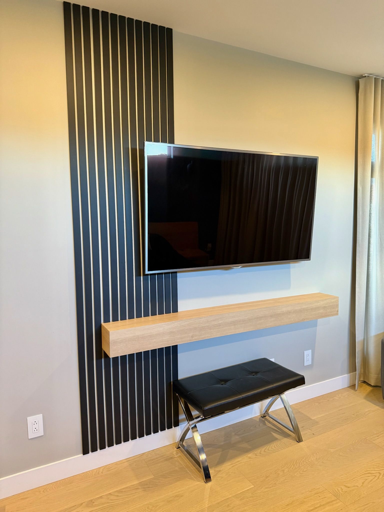 Téléviseur fixé sur un mur gris avec des panneaux de bois noirs, une étagère en bois clair et un banc noir.