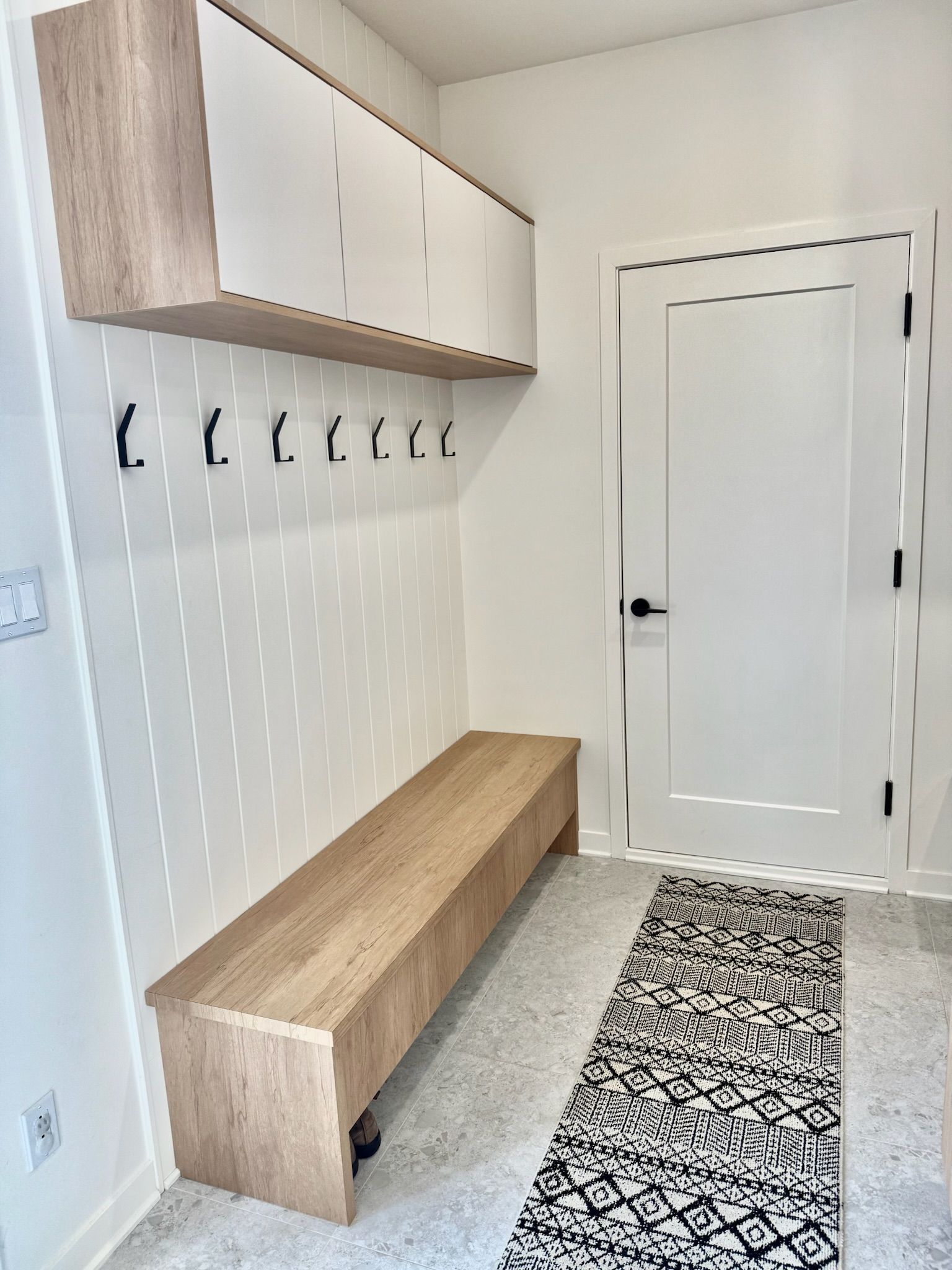 Vestibule avec banc, patères, placards suspendus et porte. Bois clair et touches de blanc.