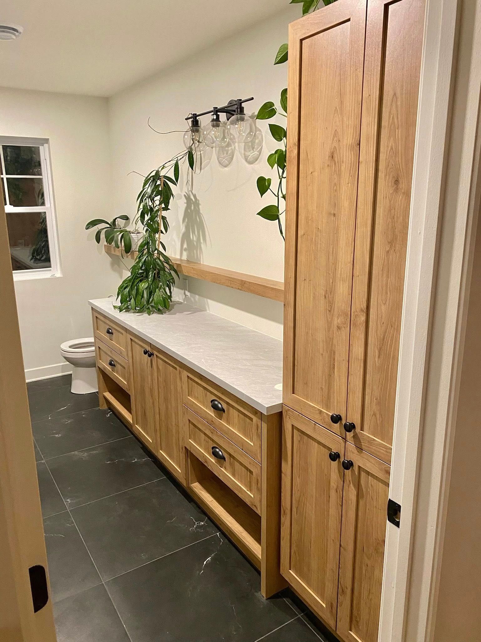 Salle de bains avec meubles en bois, plan de travail clair, carrelage foncé au sol et fenêtre. Des plantes en pot apportent une touche de verdure.