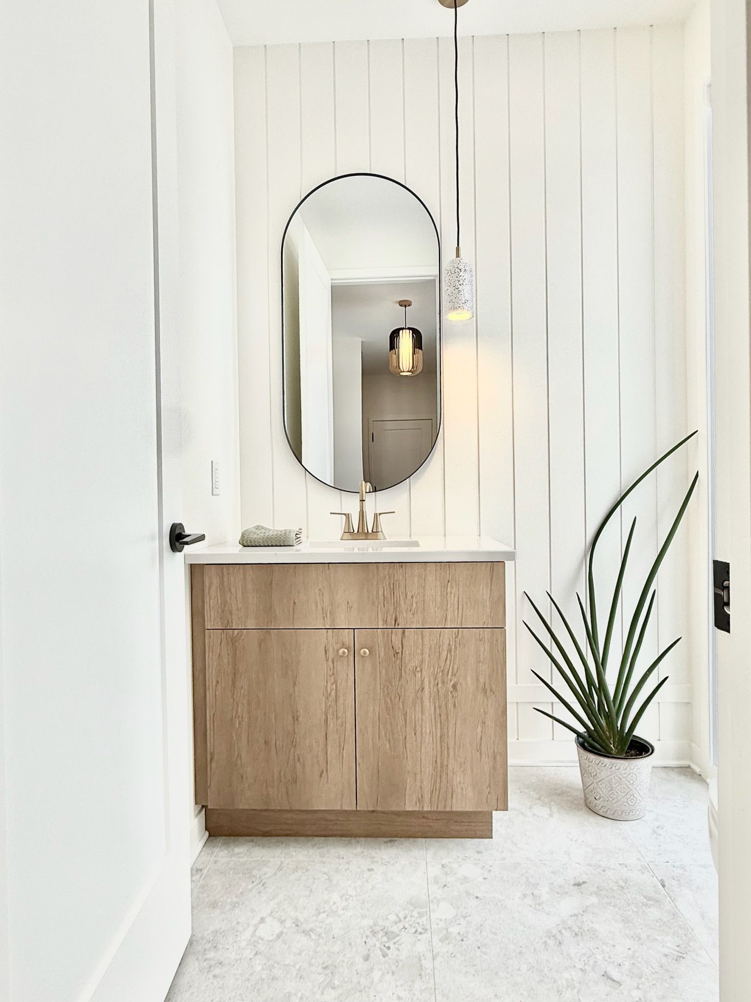 Salle de bains avec meuble-lavabo en bois, miroir ovale, lambris blanc, suspension lumineuse et plante en pot.