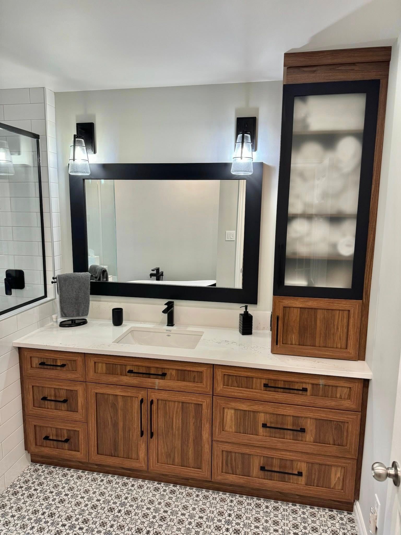 Salle de bains avec meuble-lavabo en bois, robinetterie noire, miroir et armoire. Plan de travail et murs blancs, sol en galets.