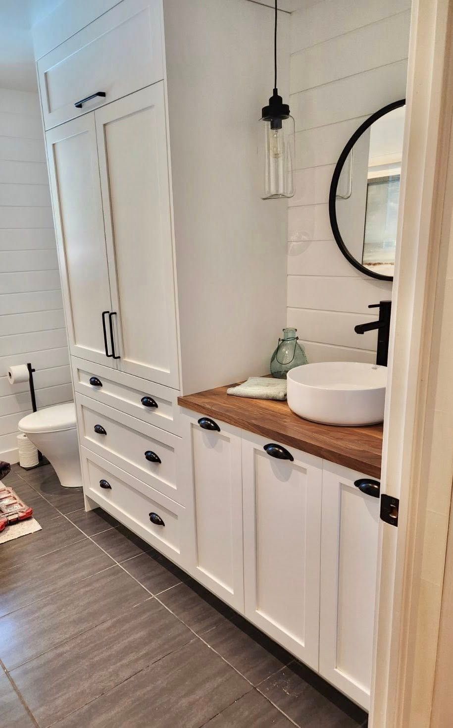 Salle de bains blanche avec rangements intégrés, un miroir rond et un plan de travail en bois avec lavabo.