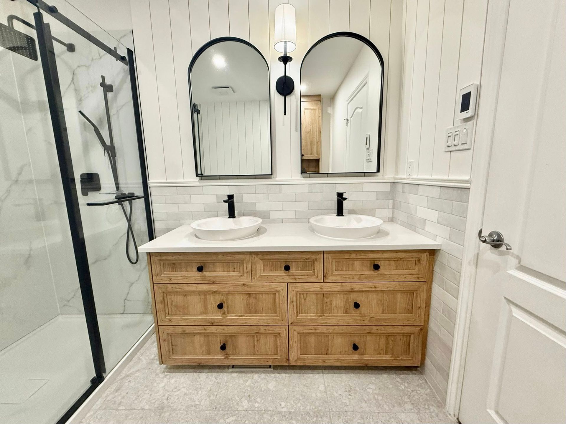 Salle de bains avec meuble-lavabo en bois, deux lavabos blancs, miroirs arqués et douche à cadre noir.