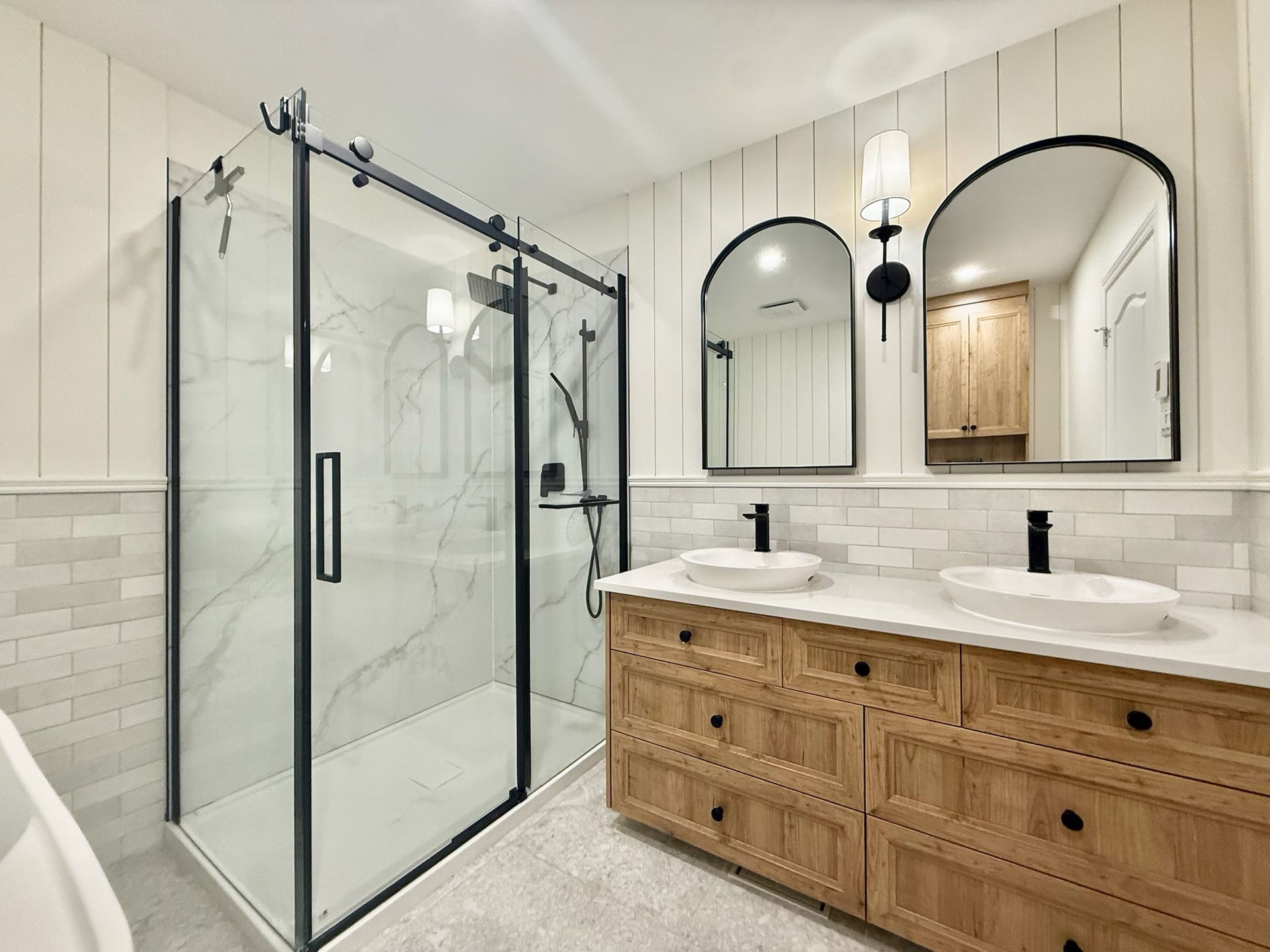 Salle de bains moderne avec douche en verre, double vasque, miroirs arqués et meubles en bois.