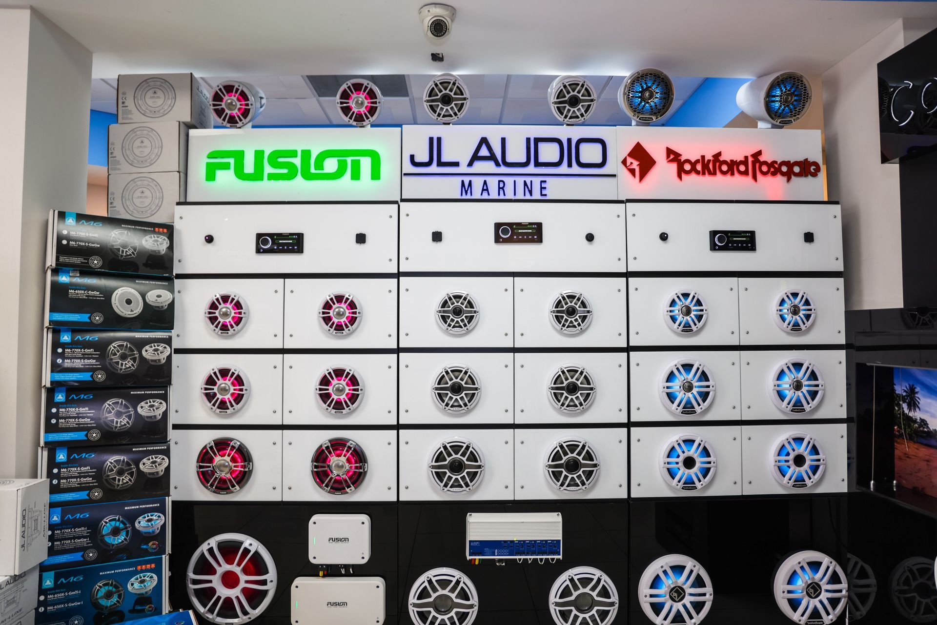 Marine audio equipment display featuring speakers, amplifiers, and branded signs: Fusion, JL Audio, and Rockford Fosgate.