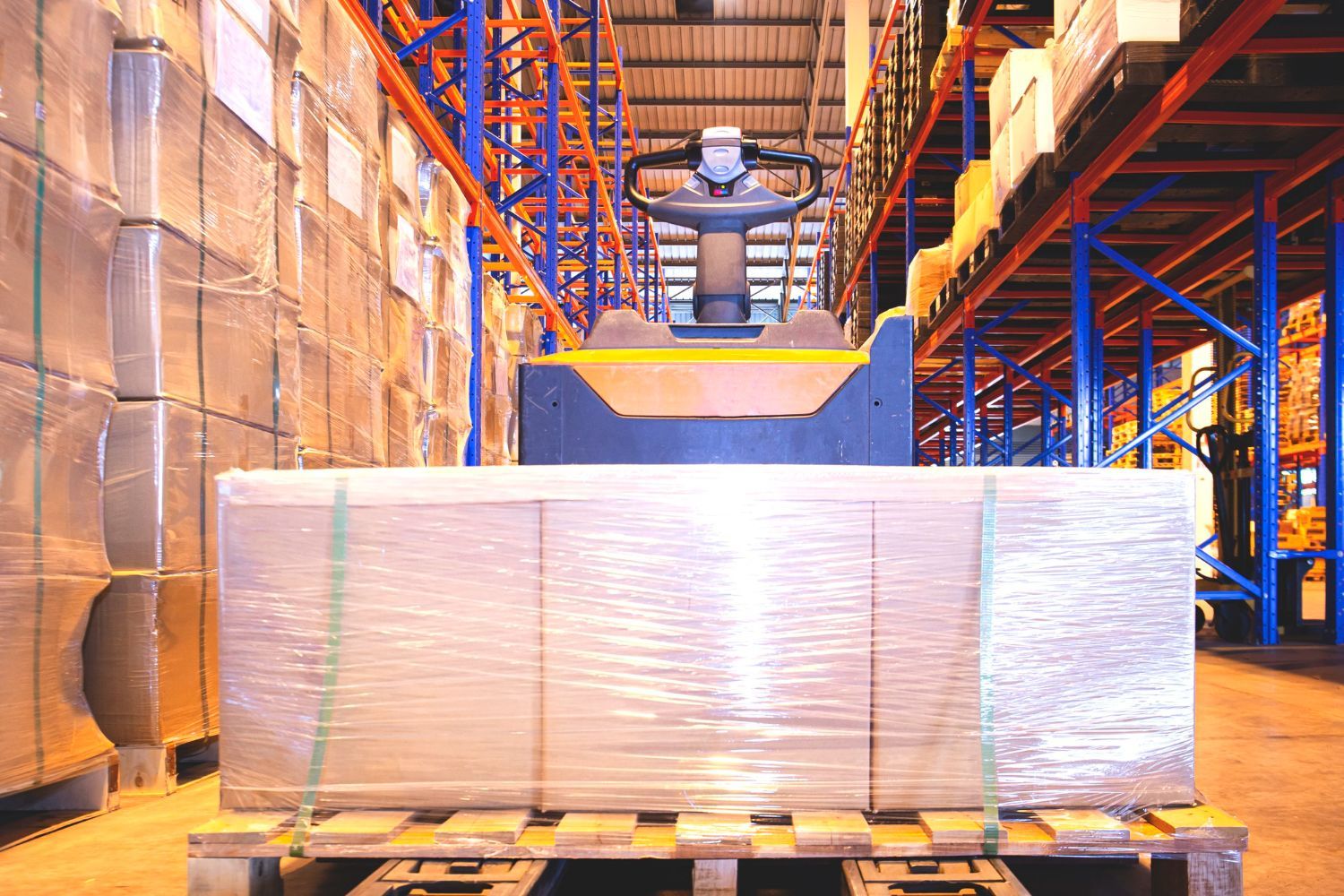 Forklift carrying wrapped pallet of goods in a warehouse aisle with shelving.