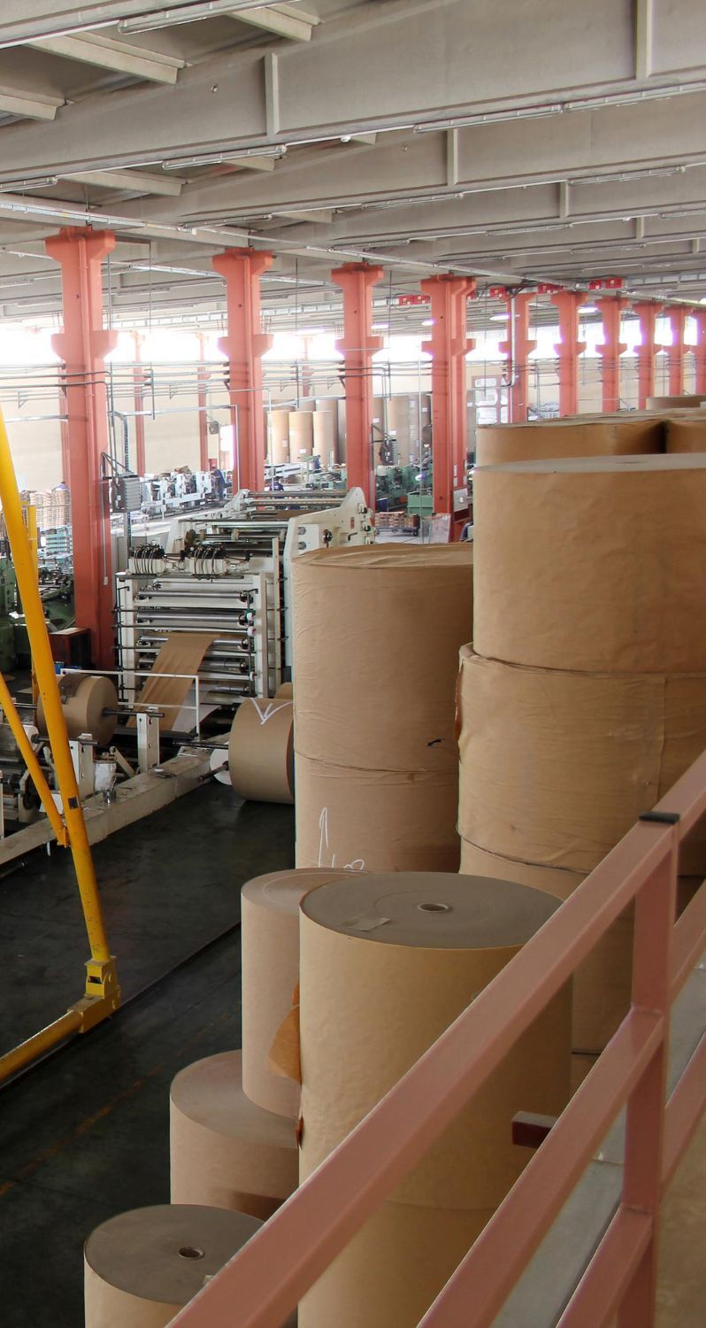 Paper rolls stacked in a warehouse. Red support beams line the ceiling, machinery in the background.