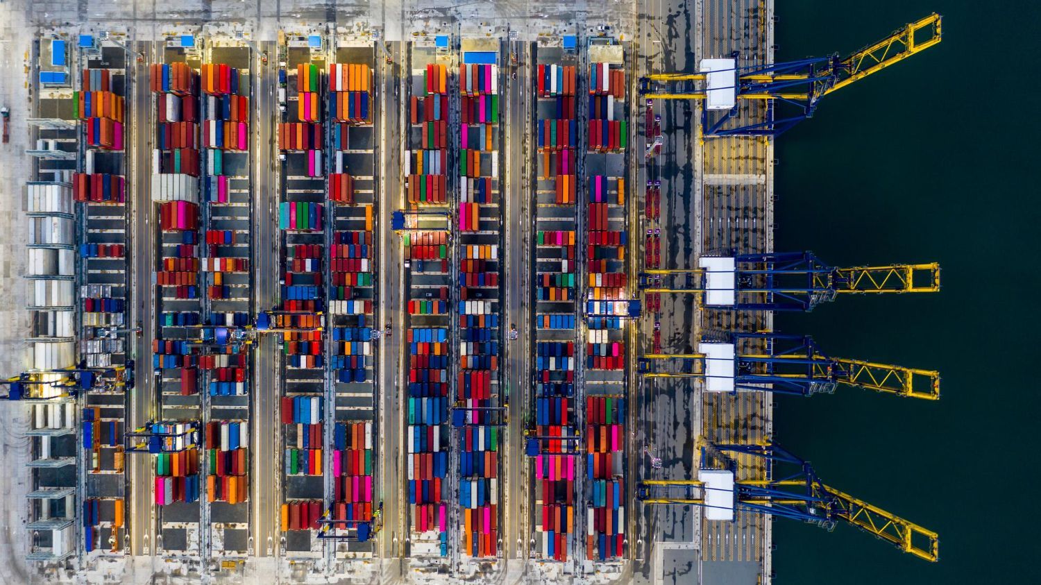 Aerial view of a busy shipping port. Colorful cargo containers are stacked neatly, with large cranes alongside a body of water.