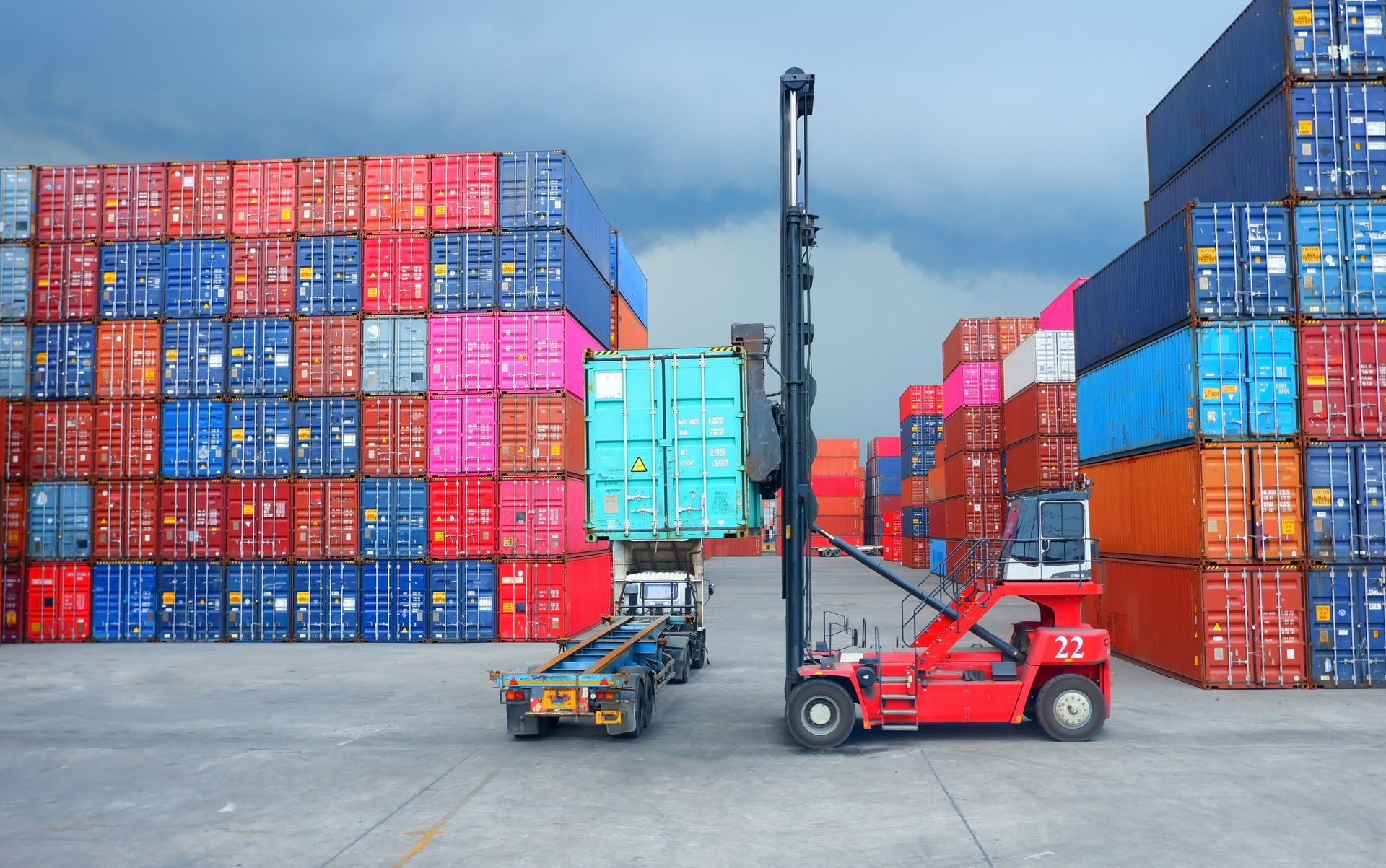 Forklift lifting a teal shipping container at a port, surrounded by stacks of colorful cargo containers.