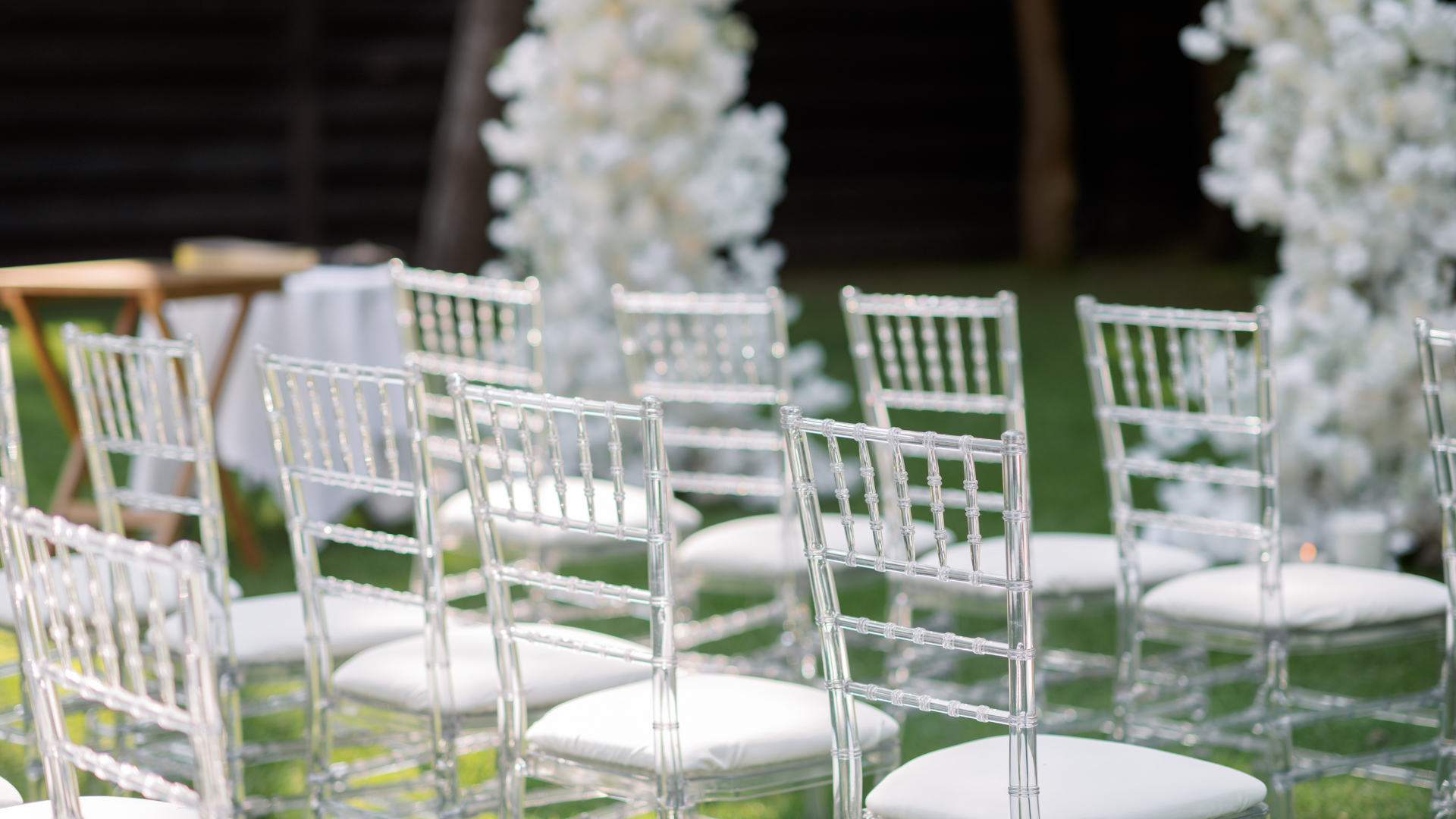 Rows of clear acrylic chairs set up for an outdoor event, with white floral decorations in the background.
