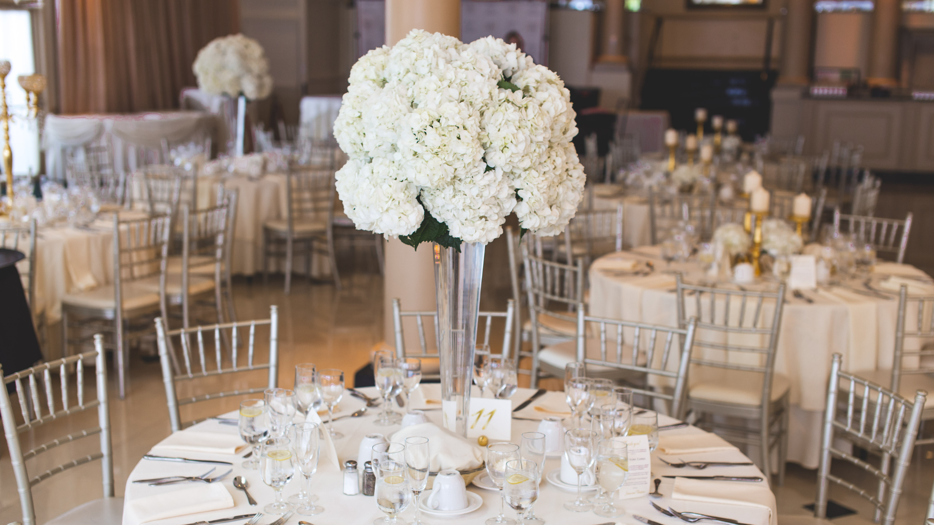 Elegant reception tables with white floral centerpieces. Silver chairs, cream tablecloths, and a formal indoor setting.