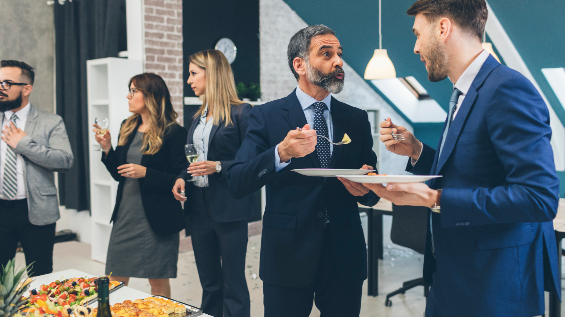 Business people in suits converse at an office event with food and drinks.