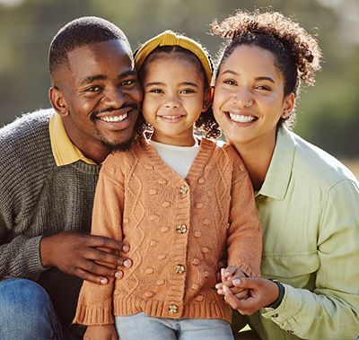 Family portrait: smiling parents flank a young girl, outdoors. The girl wears a sweater, and the parents are joyful.
