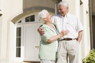 An elderly couple smiles and embraces in front of a white door. The man has his arm around the woman; they appear happy.