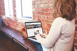 Woman using laptop with webpage open, sitting against a brick wall near a window, legs resting on a bench.