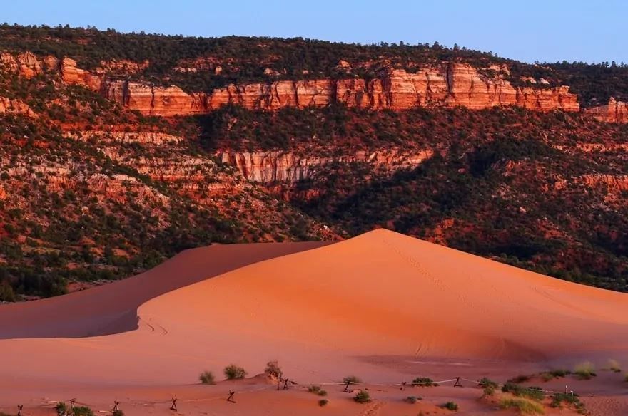 A large, orange sand dune sits at the base of a reddish-brown cliff face under a soft blue sky.
