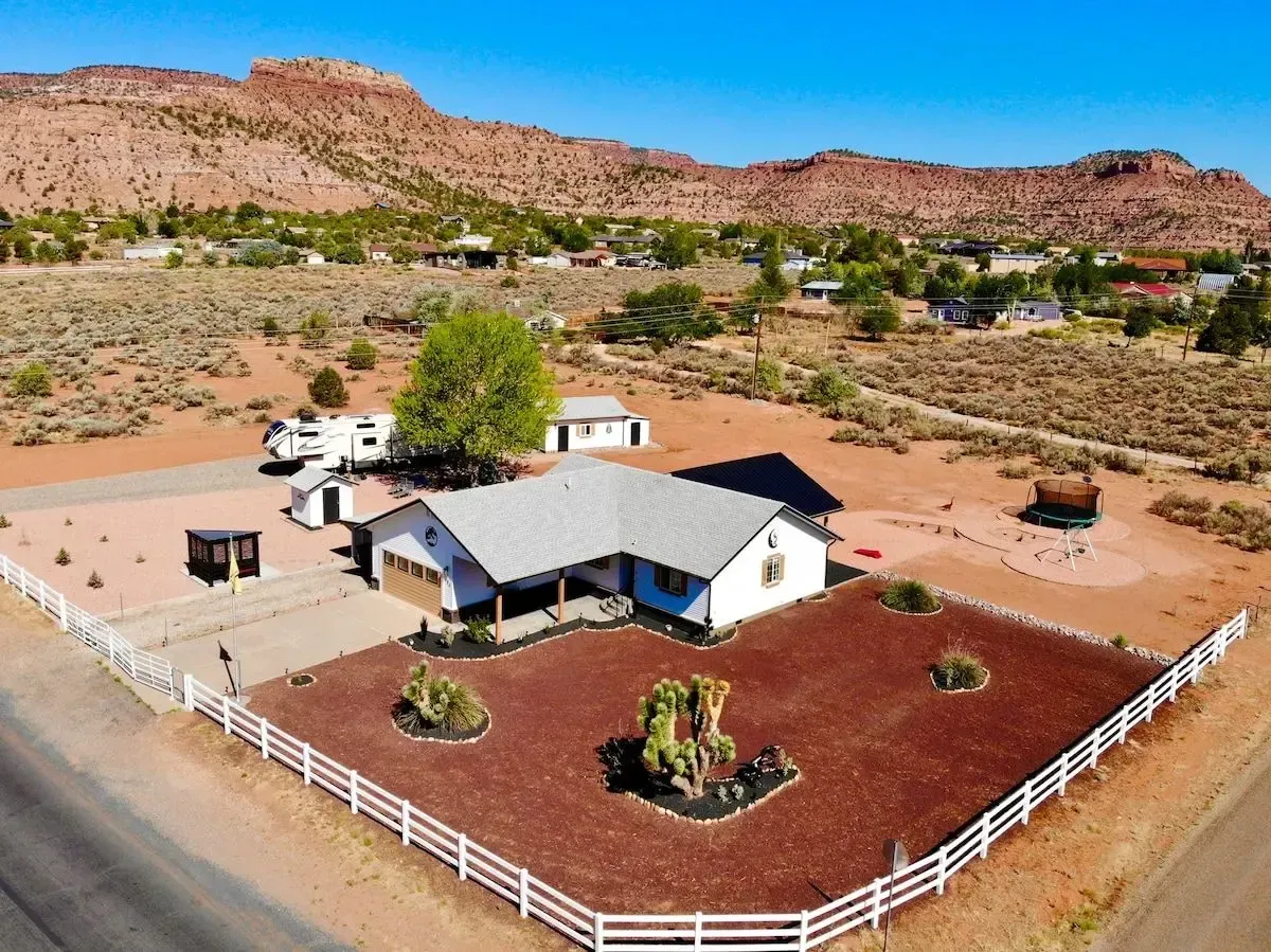 Ranch house with white fence in a desert landscape, backed by red rocky hills under a blue sky
