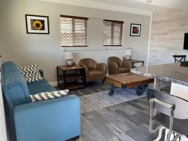 Living room with blue sofa, two tan chairs, coffee table, wall art, and windows with blinds