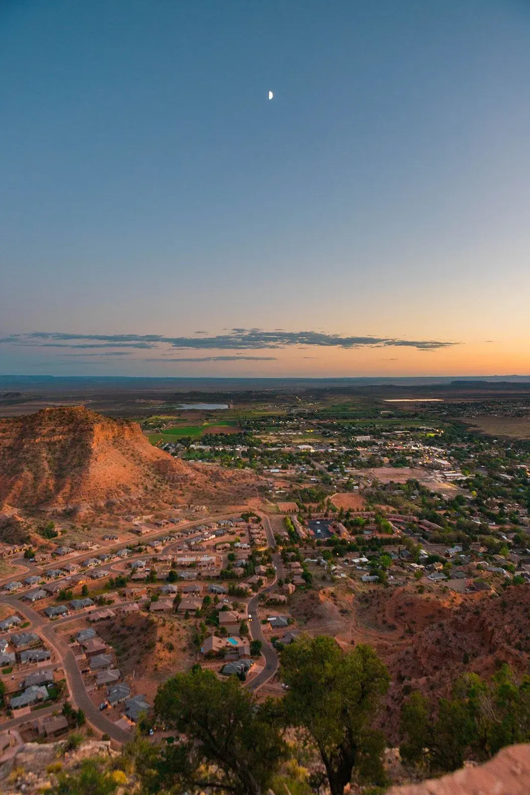 A view of a city from a cliff at sunset with a moon in the sky.