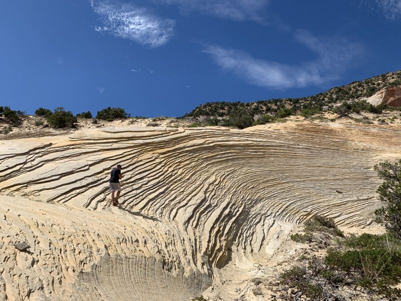 Person standing on eroded sandstone with linear patterns; blue sky and hillside in background.