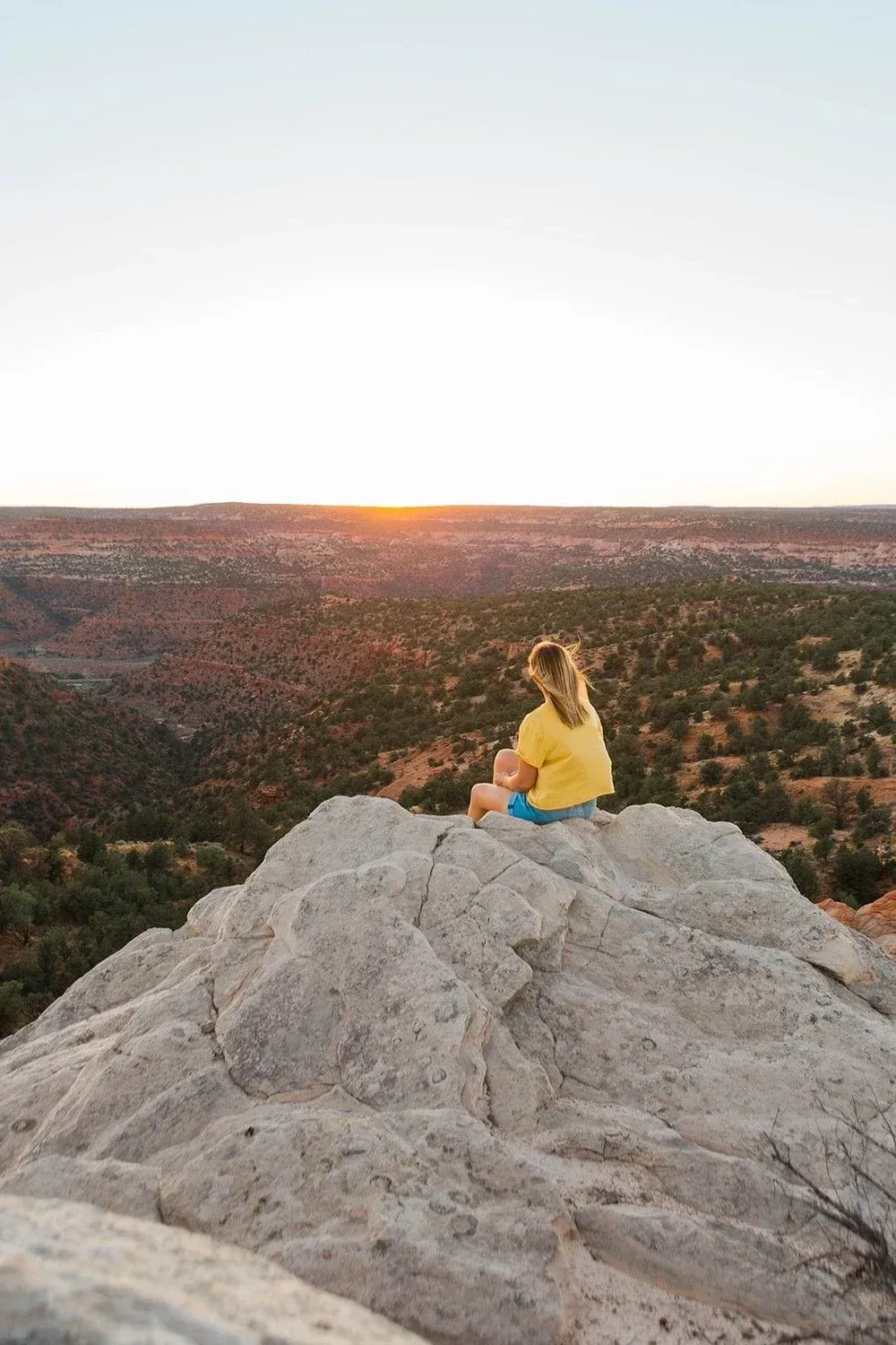 Person in a yellow top sits on a rocky outcrop at sunset overlooking a city.