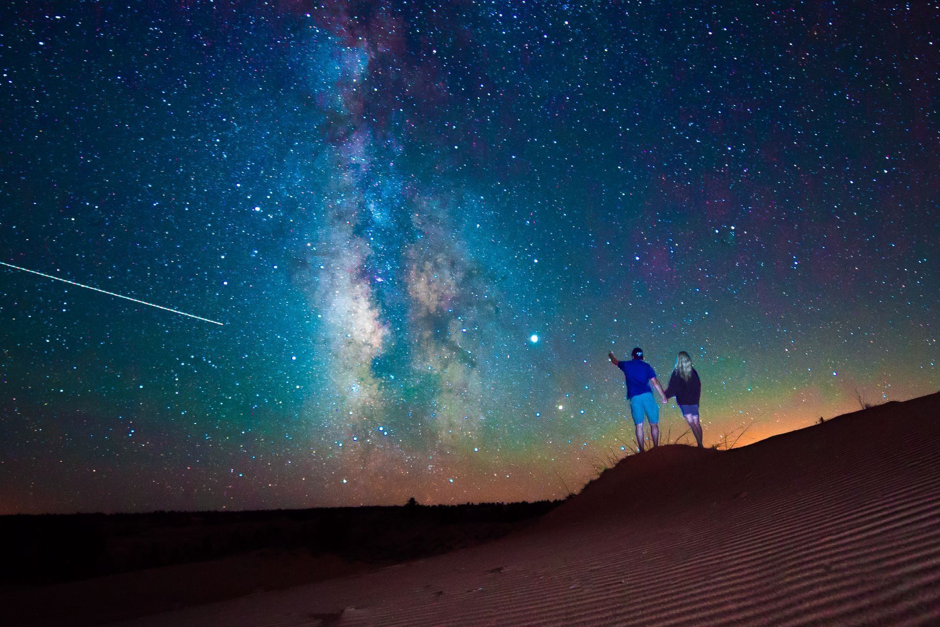 Two people are standing on top of a sand dune looking at the stars.