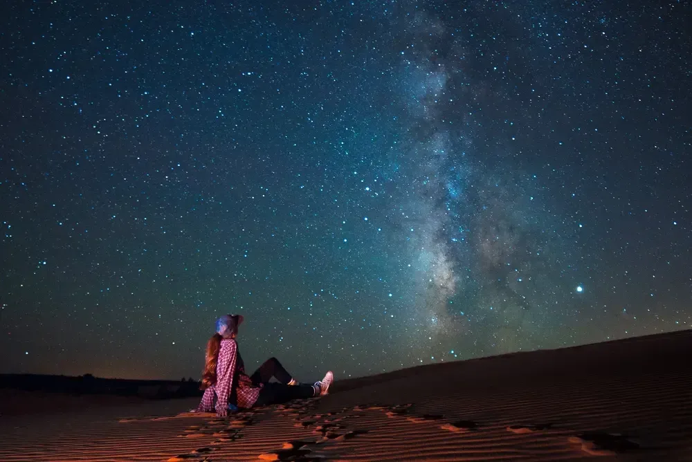 Two people on sand dunes under a starry night sky with the Milky Way overhead