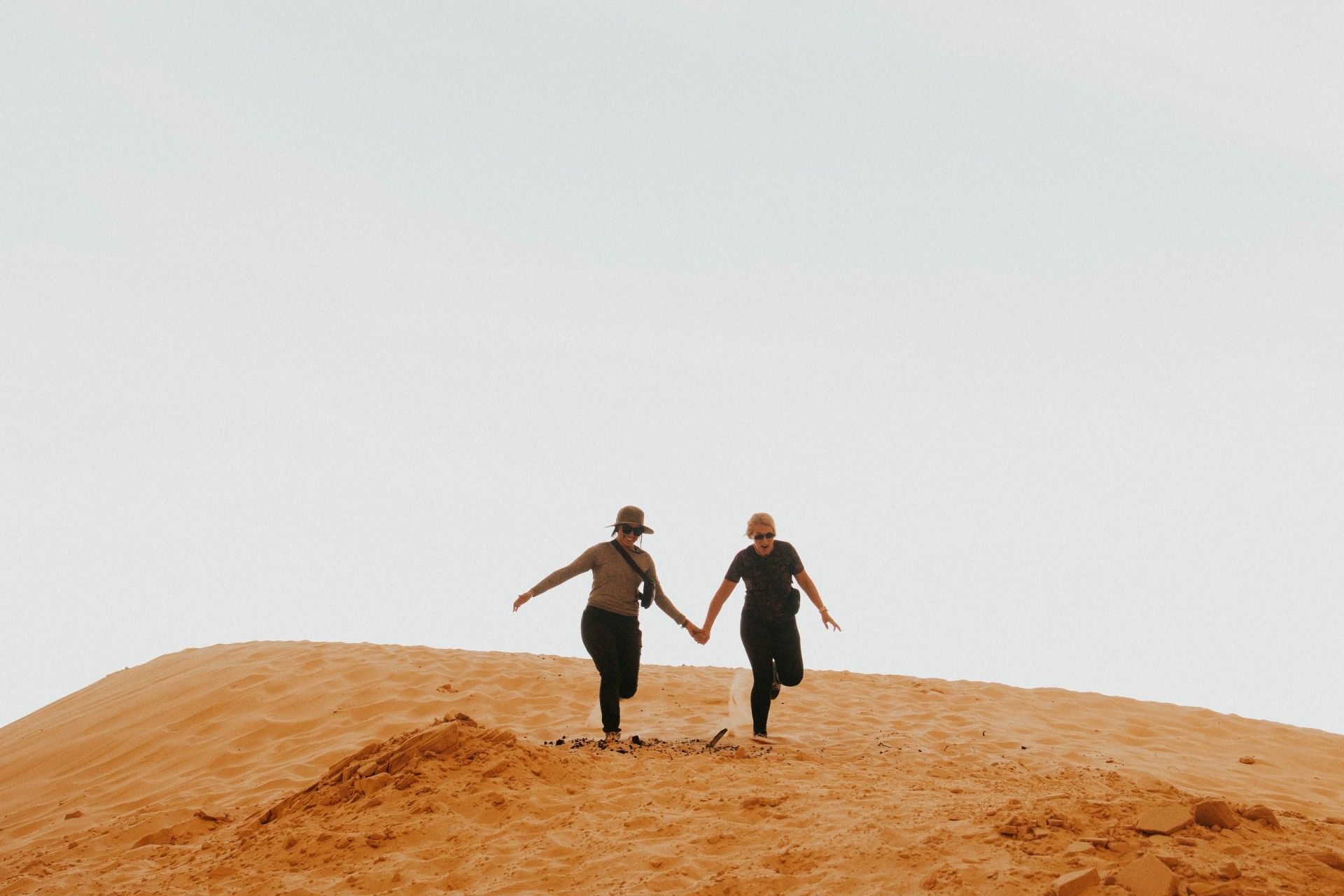 Two people are holding hands while walking on top of a sand dune.