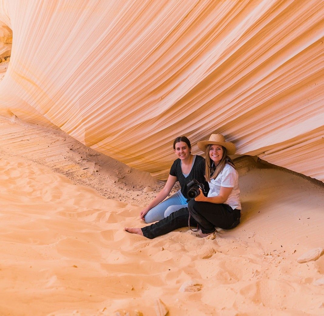 Two people sitting in a sandstone canyon. One wears a hat. The canyon walls are striped with beige and orange hues.