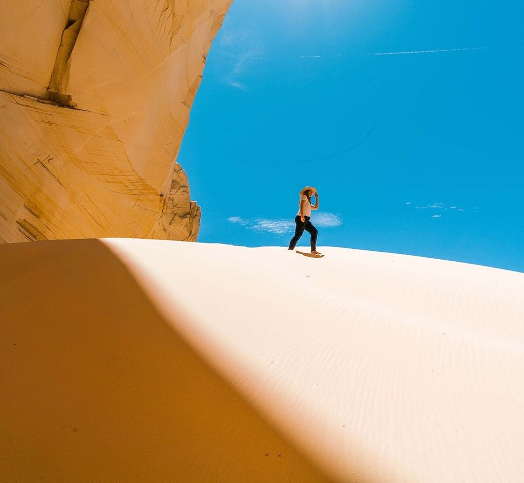 Person walking on a sand dune under a blue sky, near a sandstone cliff.