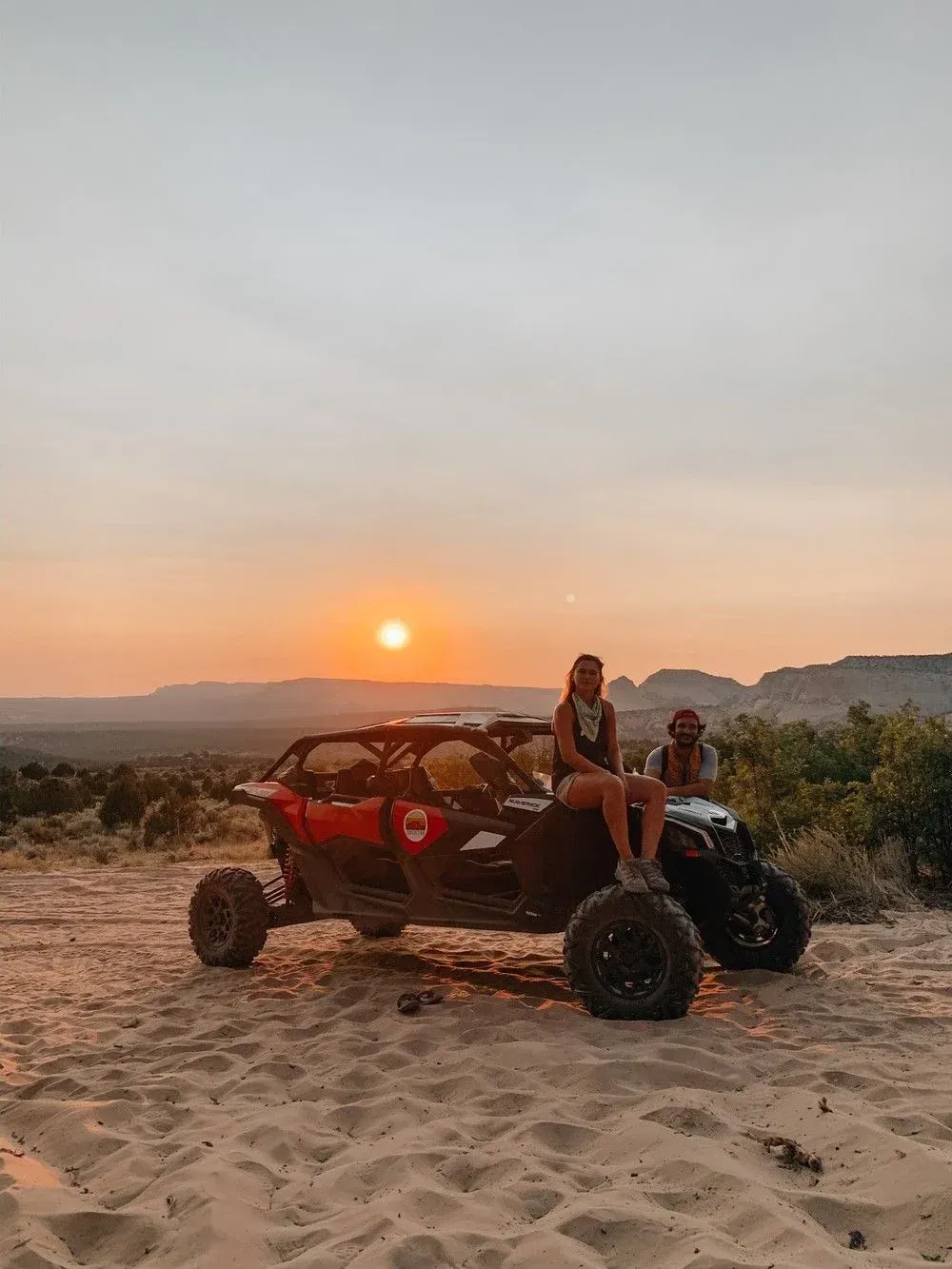 Two people on a red dune buggy at sunset on sandy terrain with mountains in the distance