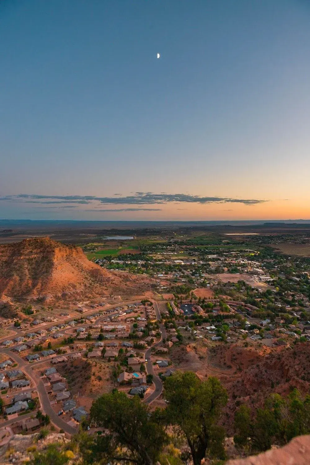 Sunset over a desert town and red rock cliffs with a crescent moon in the sky