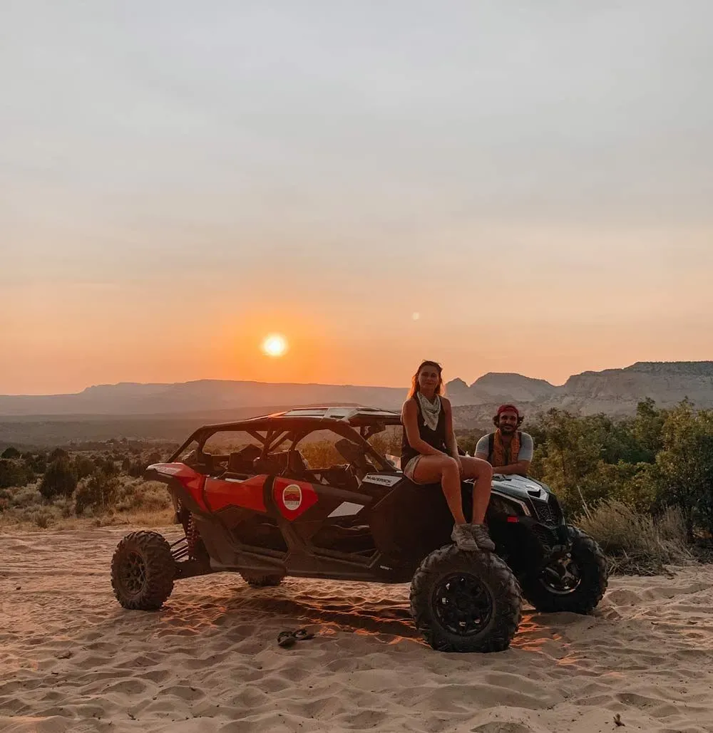 Two people are sitting on top of a atv in the desert at sunset.