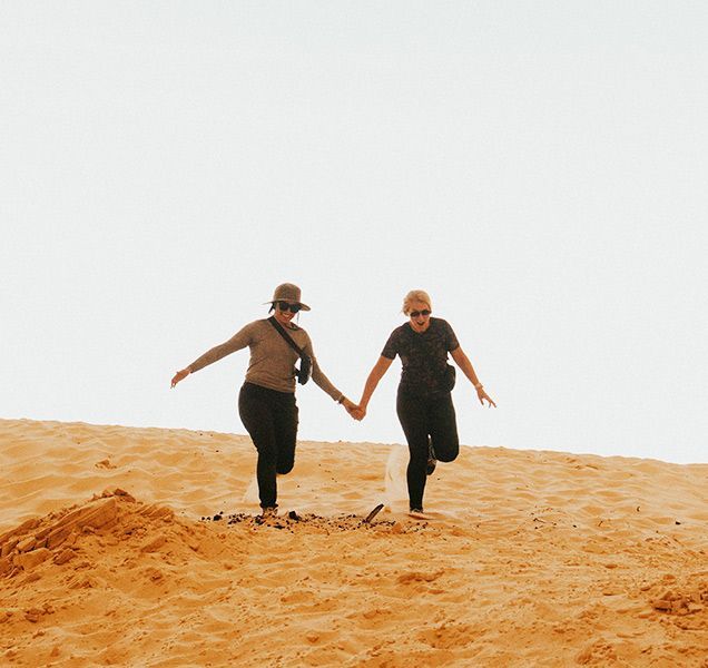 Two women are holding hands while walking on a sand dune.