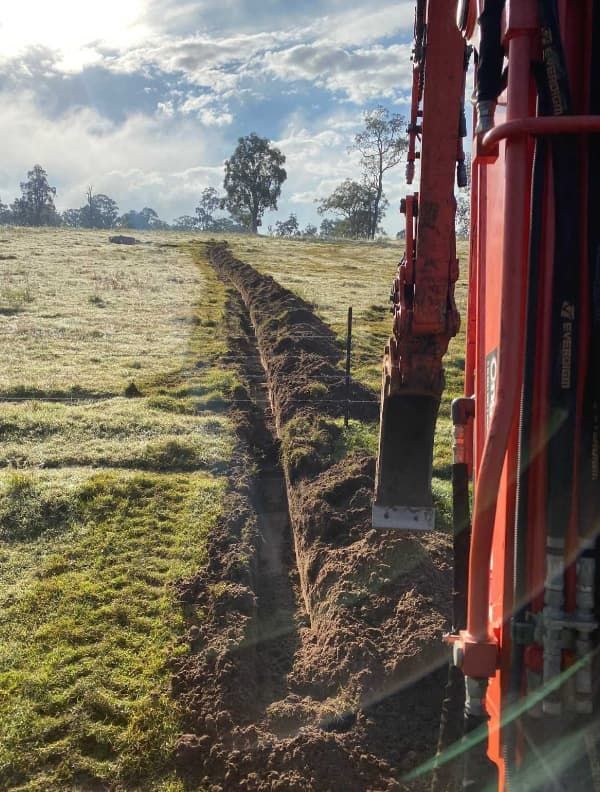 A Red Excavator Is Digging A Trench In A Grassy Field — Northstar Excavations In Alstonville, NSW