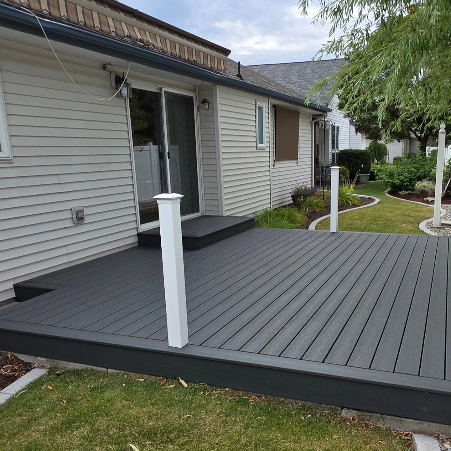 Newly built gray deck with white posts, next to a house with a sliding glass door and green lawn.