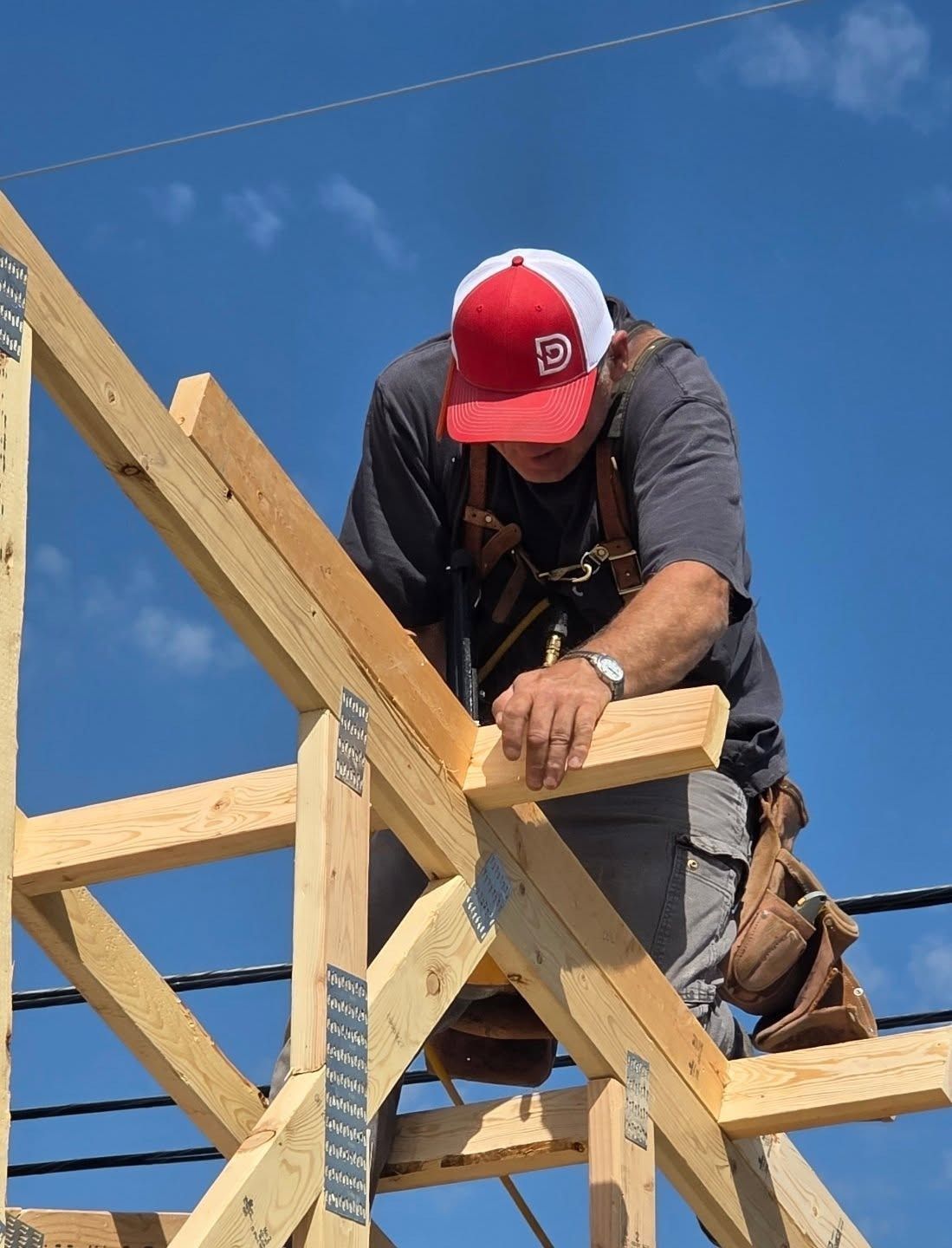 Carpenter on a roof, securing wooden beams with metal connectors. Wearing a hat and safety harness, blue sky background.