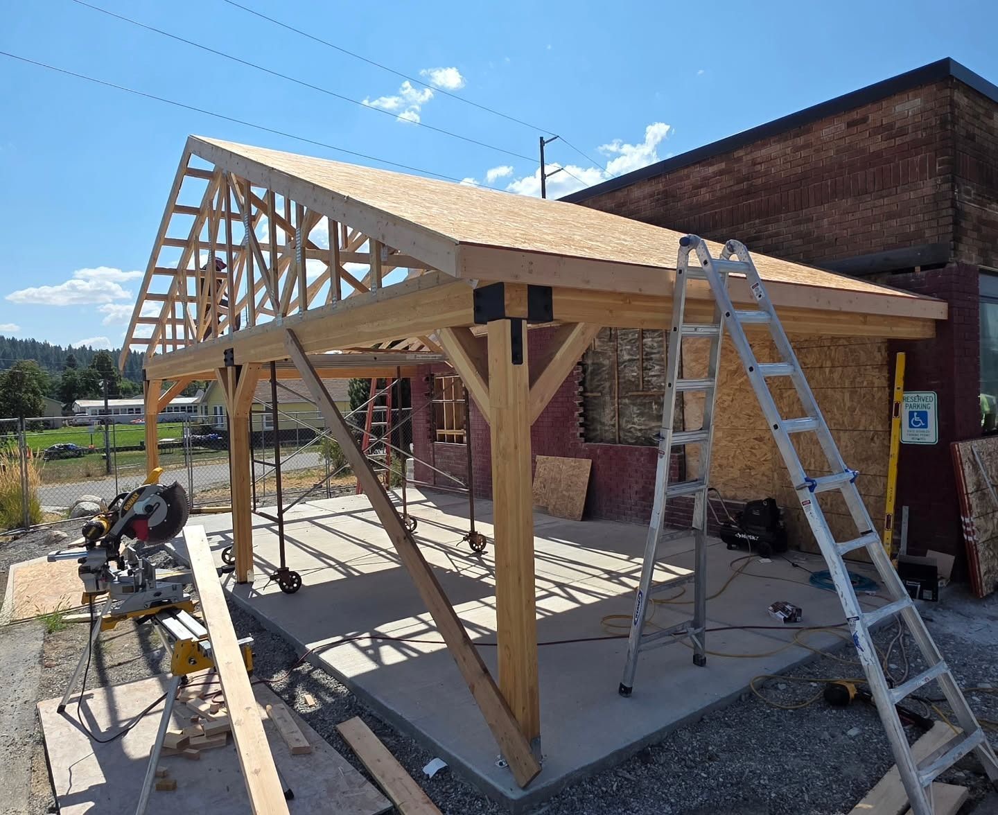 Wooden structure being built; roof frame and support beams in progress. Ladder and tools visible.
